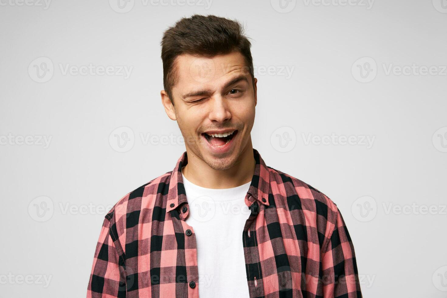 sorrindo amplamente bonito atraente encantador jovem homem, piscadelas com 1 olho, paquera sorriso, agradável aparência, vestido dentro xadrez camisa, em pé contra branco fundo. foto