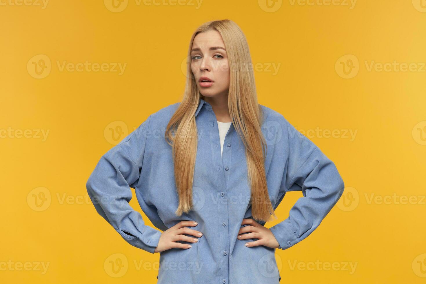 retrato do sério mulher com loiro grandes cabelo. vestindo azul camisa. pessoas e emoção conceito. coloca mãos em uma cintura. fez você dizer alguma coisa assistindo às a Câmera, isolado sobre laranja fundo foto