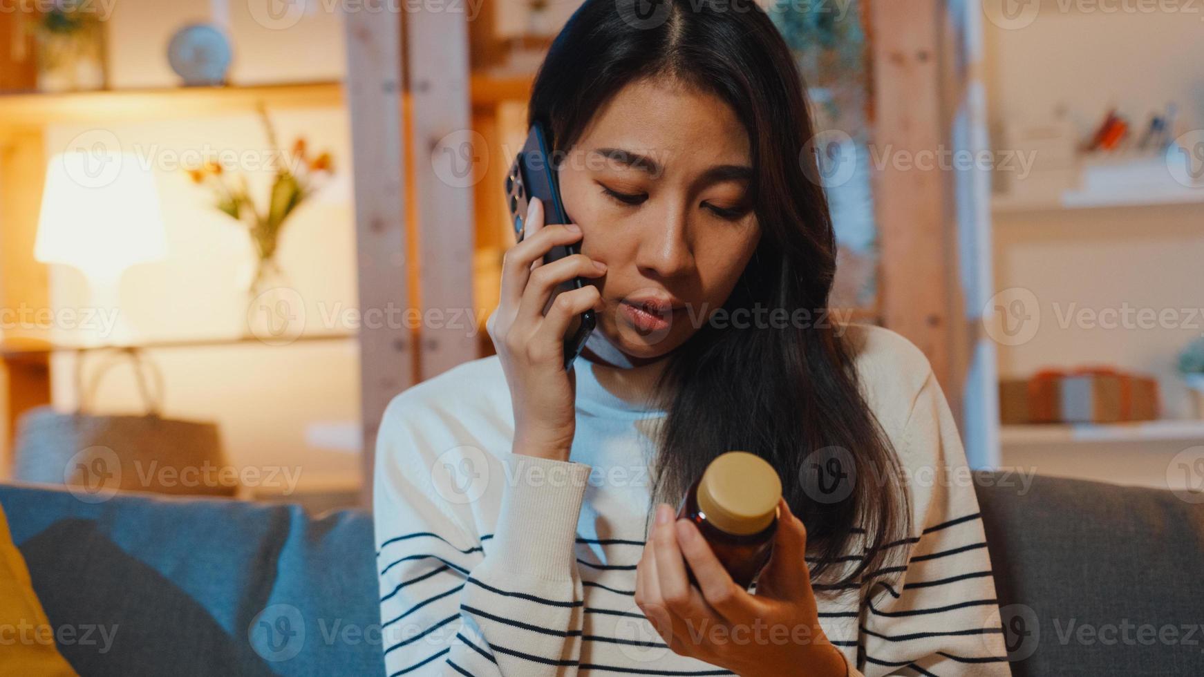 mulher asiática jovem doente segurar remédio sentar no sofá usar telefone inteligente para consultar o médico em casa à noite. garota tomar remédio após ordem do médico, quarentena em casa, conceito de coronavírus de distância social. foto