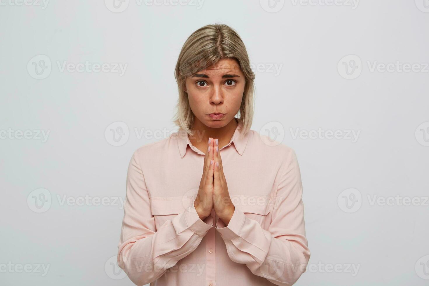 retrato do triste fofa mulher com Loiras cabelo desgasta Rosa camisa sente chateado e mantém mãos dentro Rezar posição isolado sobre branco fundo parece diretamente dentro Câmera foto