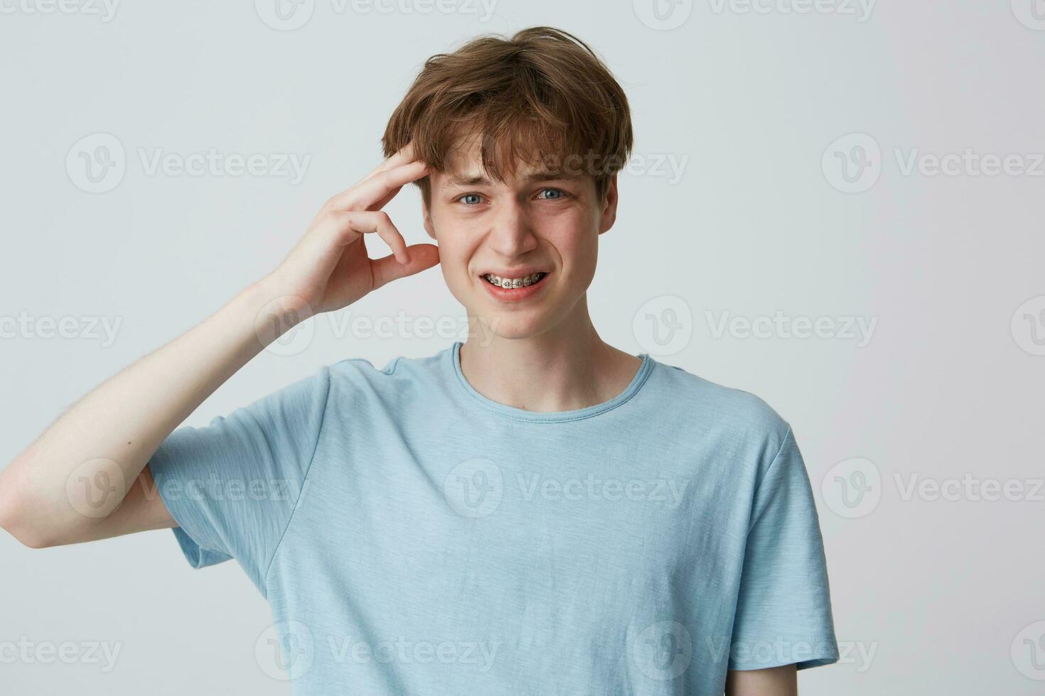 retrato do chateado deprimido jovem homem com suspensórios em dentes desgasta azul t camisa tocante dele têmpora e ter uma dor de cabeça isolado sobre branco fundo foto