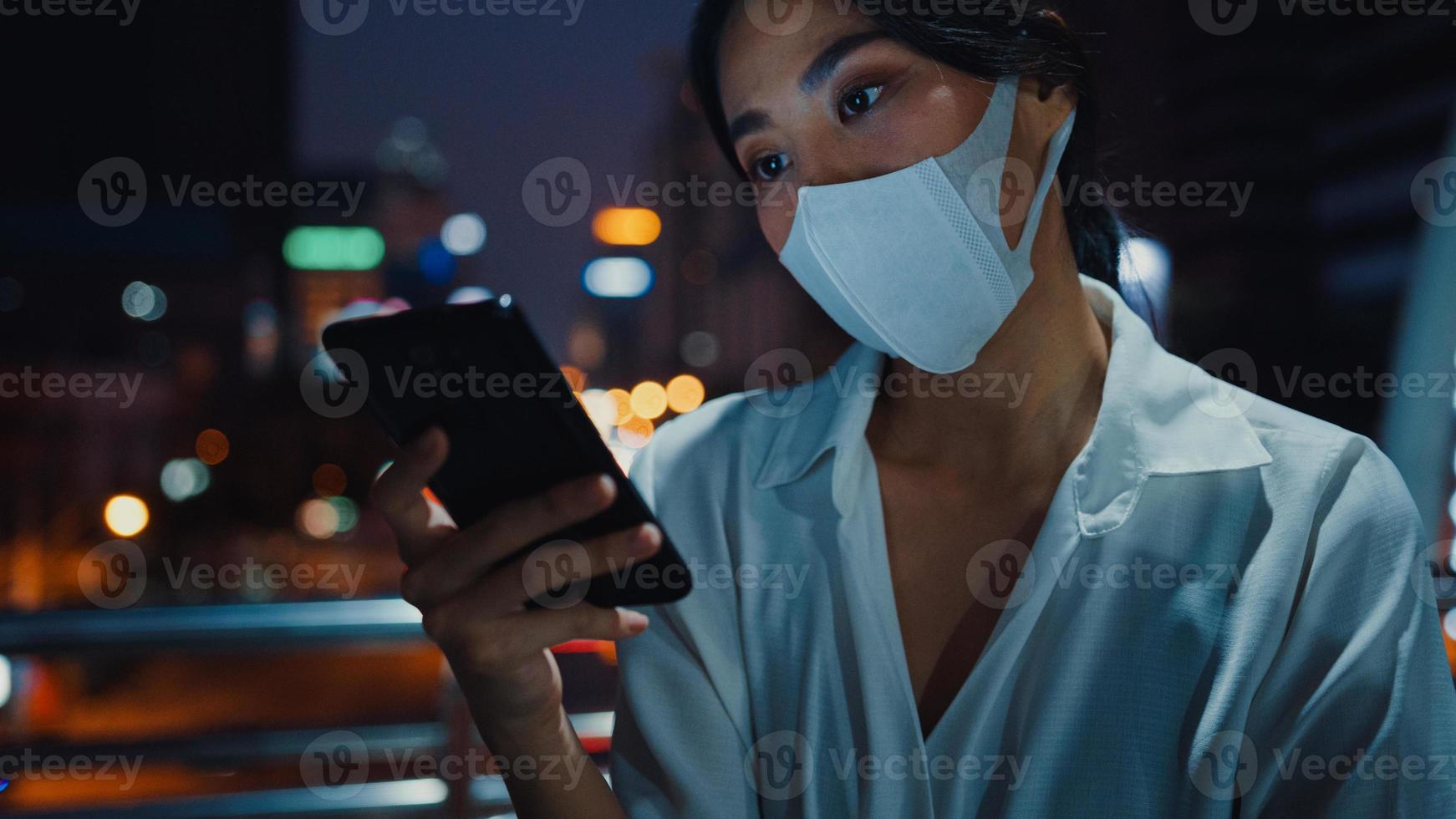 jovem empresária asiática em roupas da moda, usando máscara facial, usando telefone inteligente, digitando mensagem de texto, enquanto fica ao ar livre na cidade urbana à noite. distanciamento social para evitar a disseminação do conceito covid-19. foto