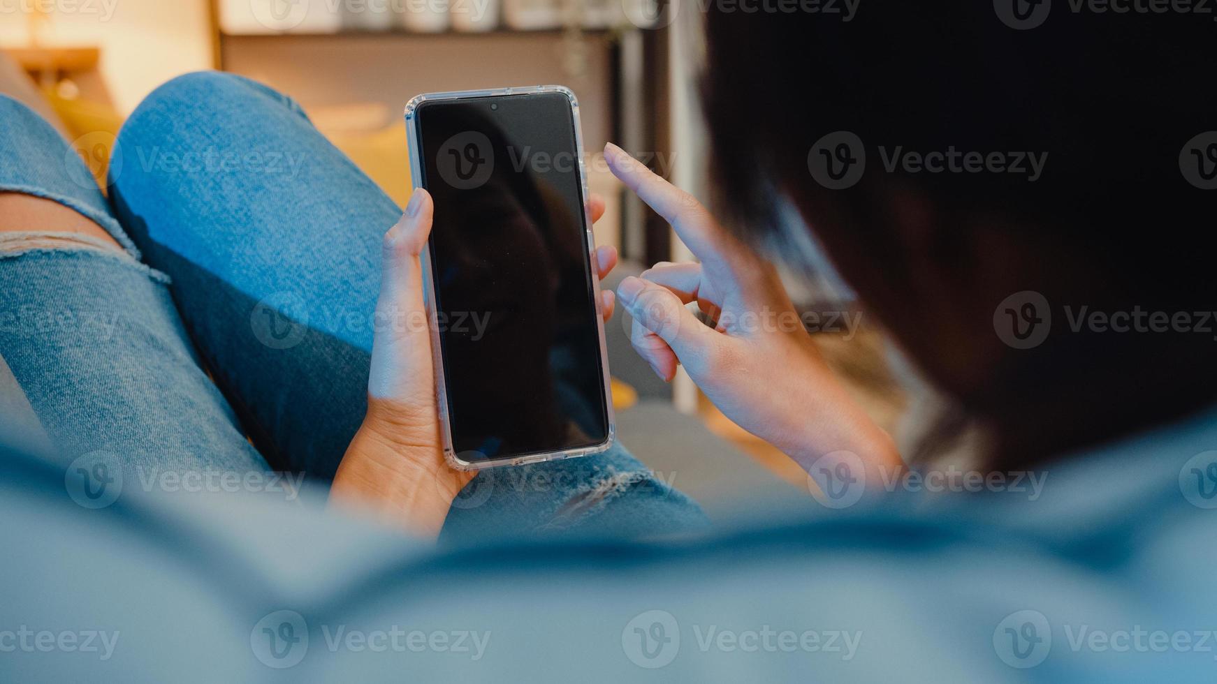 jovem asiática usar telefone inteligente com simulação de tela preta em branco para texto de publicidade, enquanto descansa no sofá na sala de estar em casa à noite moderna. tecnologia chroma key, conceito de design de marketing. foto