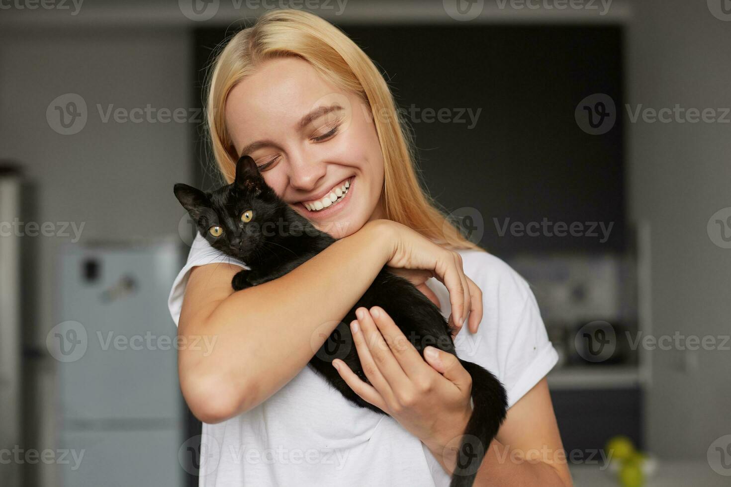 feliz jovem Loiras menina às a cozinha detém em mãos, carícias, acariciando, abraços Preto pequeno gato, fofa ternamente sorridente, olhos fechado, vestido dentro uma doméstico branco camiseta, cozinha fundo foto