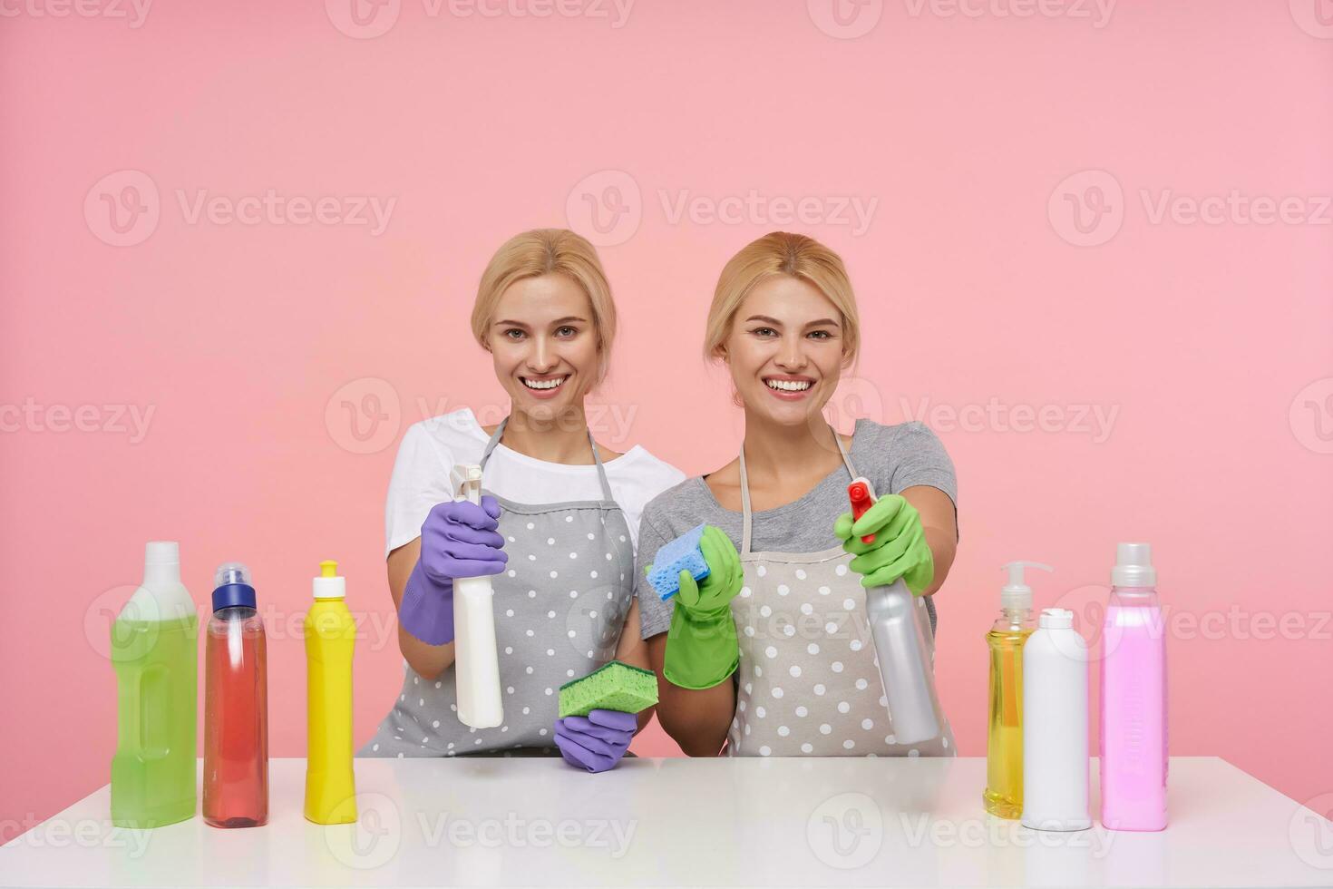 alegre jovem atraente cabeça branca limpeza senhoras levantando mãos com spray garrafas e olhando alegremente às Câmera com amplo sorrisos, sentado sobre Rosa fundo foto