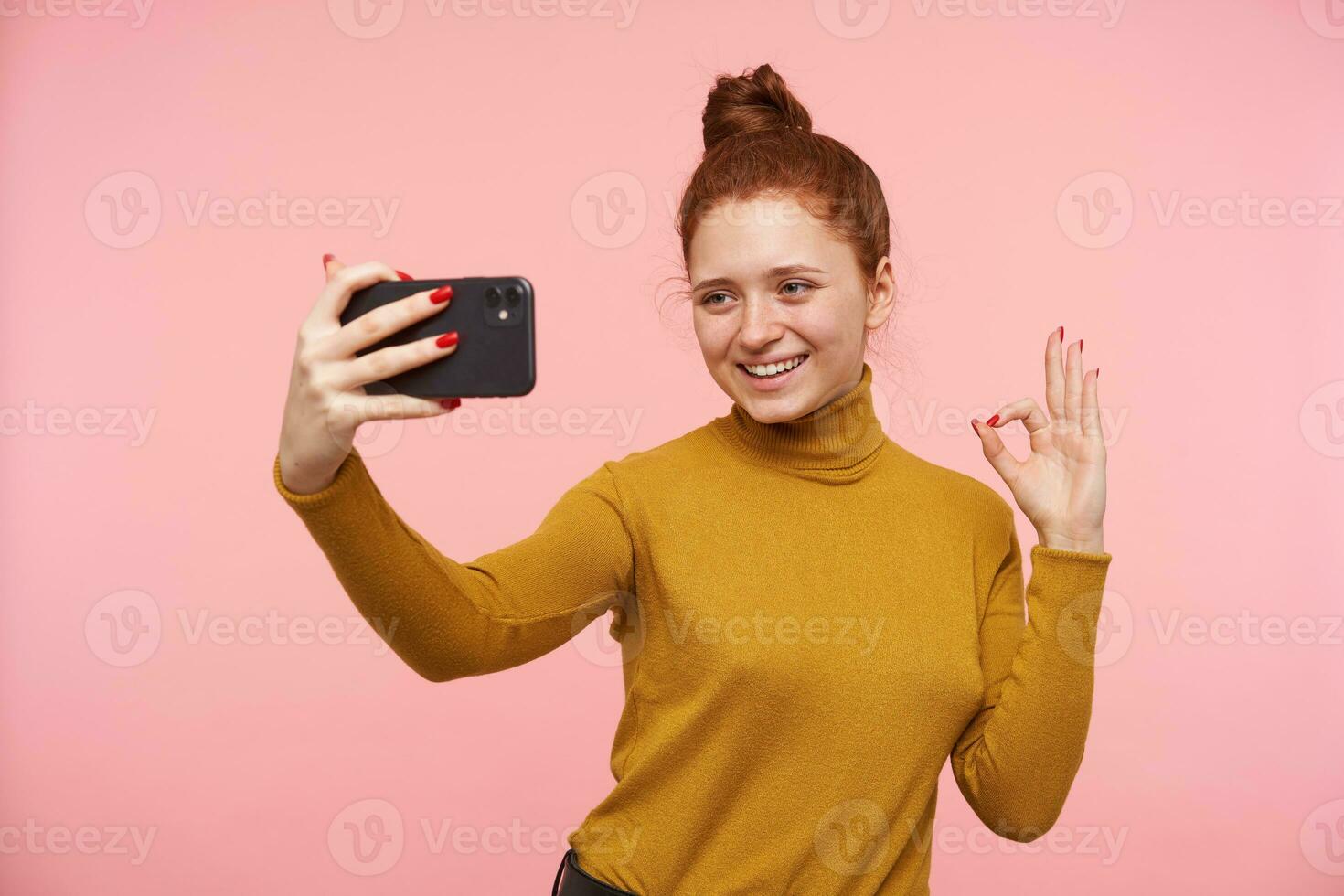 feliz jovem lindo ruiva mulher com casual Penteado levantando mão com Está bem gesto enquanto fazer selfie com dela Móvel telefone, em pé contra Rosa fundo foto