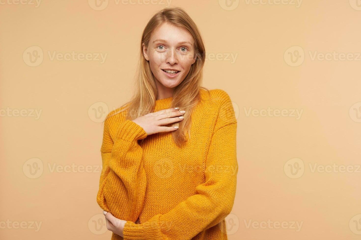 retrato do positivo jovem adorável ruiva mulher segurando elevado Palma em dela peito enquanto olhando às Câmera, vestindo acolhedor tricotado suéter enquanto posando sobre bege fundo foto