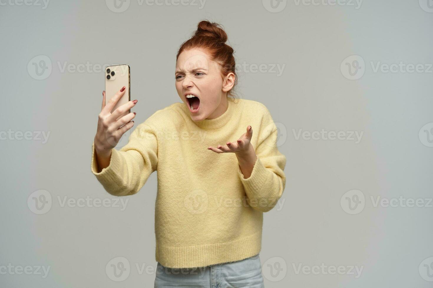 Bravo olhando mulher, gritando ruiva menina com cabelo pão. vestindo amarelo suéter e segurando uma Smartphone. discutindo com alguém em vídeo bater papo. chateado desligado. ficar de pé isolado sobre cinzento fundo foto