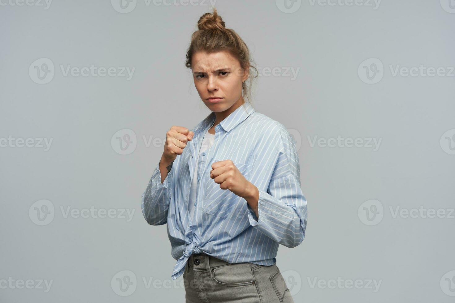 retrato do sério, adulto menina com loiro cabelo colhido dentro pão. vestindo listrado atado camisa. apertar dela punhos e pronto para lutar. assistindo às a Câmera, isolado sobre cinzento fundo foto