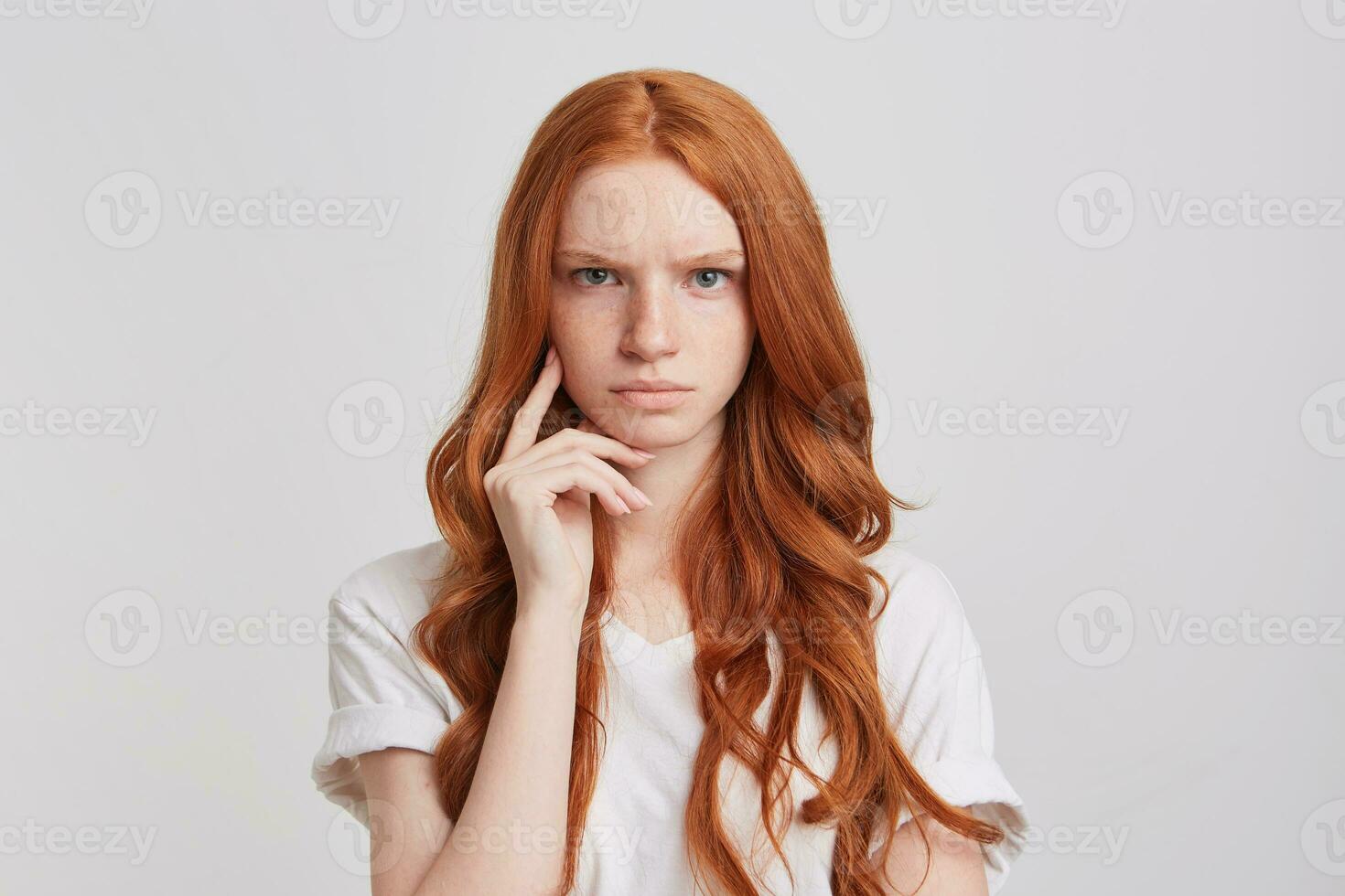 fechar-se do sério rigoroso ruiva jovem mulher com grandes ondulado cabelo desgasta t camisa parece Bravo e concentrado isolado sobre branco fundo foto