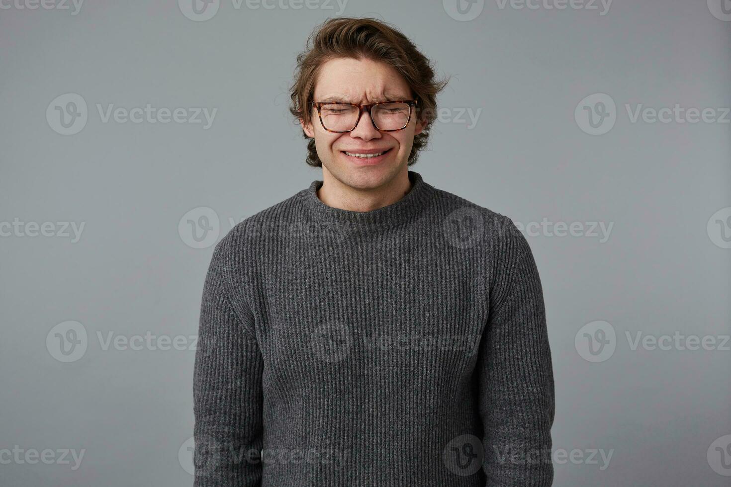 foto do jovem chorando bonito homem com óculos desgasta dentro cinzento suéter, carrinhos sobre cinzento fundo e parece infeliz e triste.