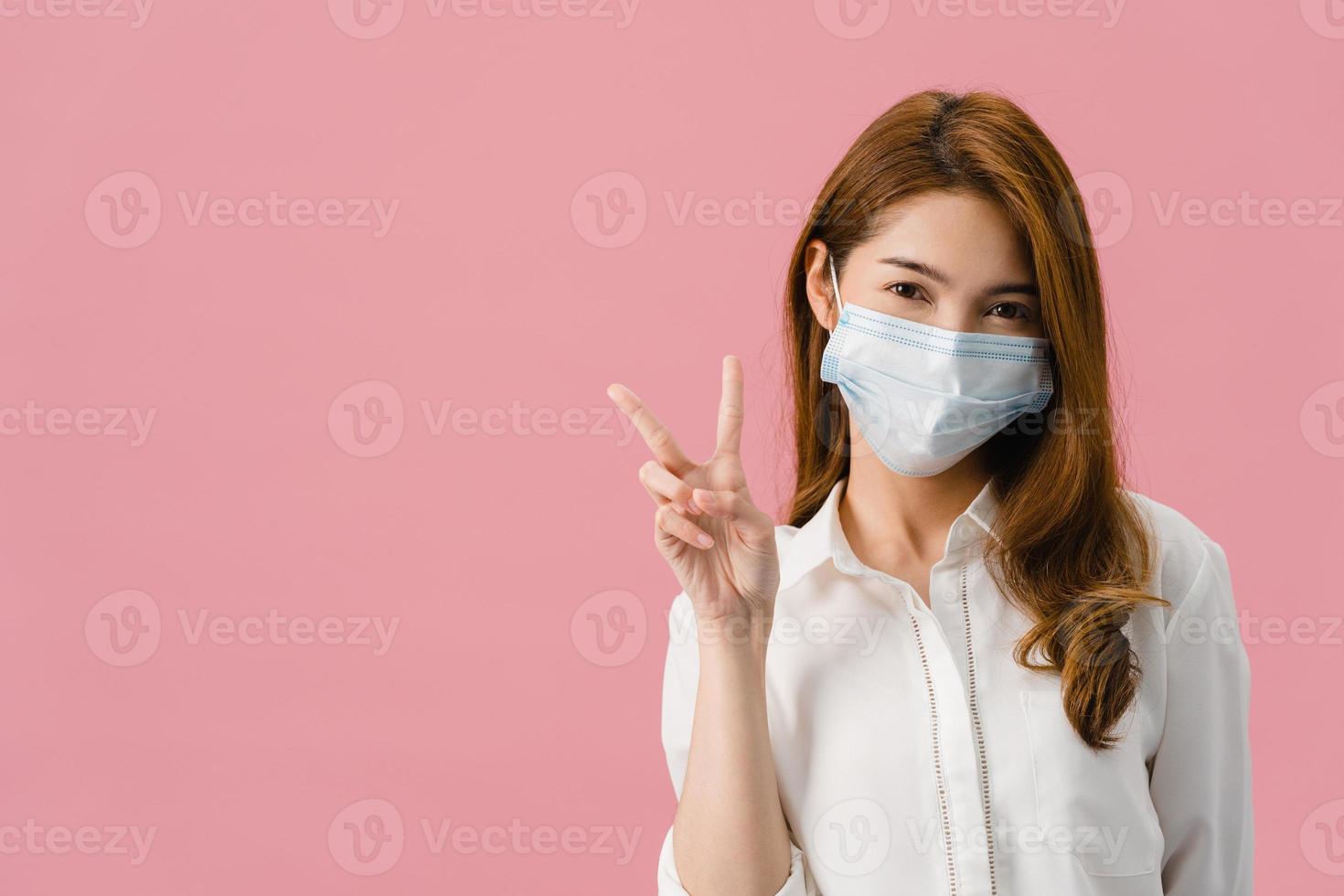 jovem asiática usando máscara médica mostrando o símbolo da paz, incentive com vestido de pano casual e olhando para a câmera isolada no fundo rosa. distanciamento social, quarentena para o vírus corona. foto