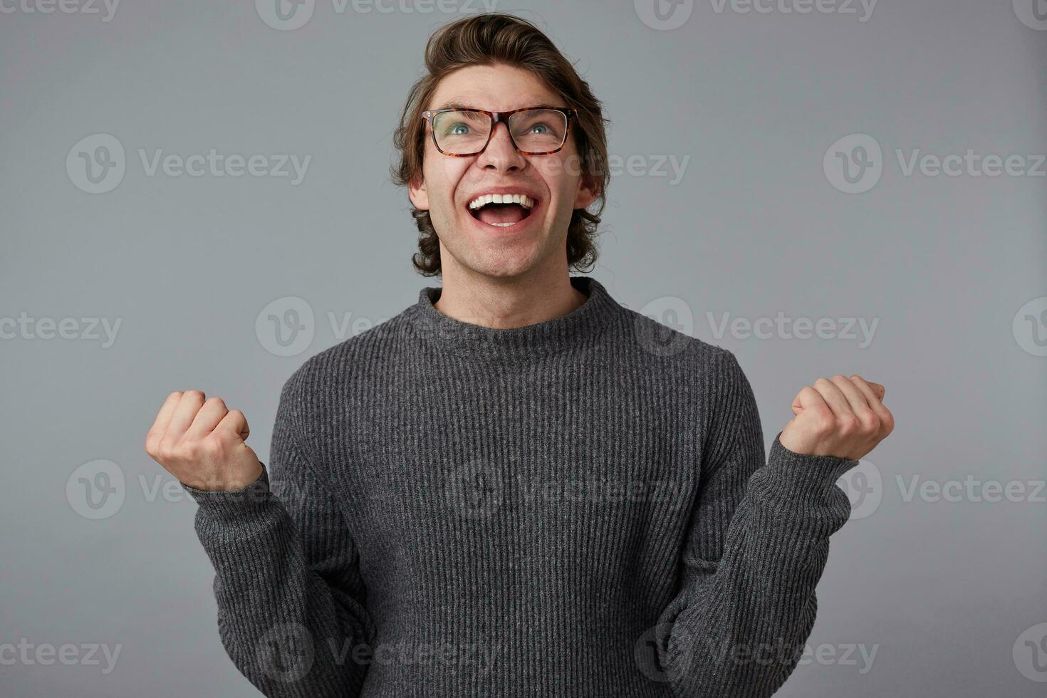 foto do jovem feliz cara com óculos desgasta dentro cinzento suéter, carrinhos sobre cinzento fundo. amplamente sorrisos e aperta punhos, Ganhou a milhão e sente felicidade.