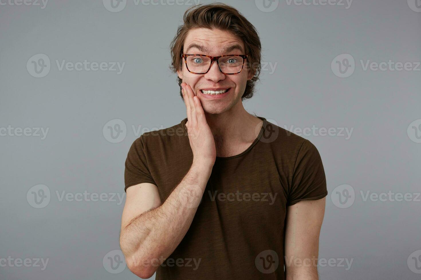 retrato do jovem bonito homem com óculos desgasta dentro básico Camisetas, carrinhos sobre cinzento fundo e amplamente sorrisos, parece feliz e perguntou-se. foto