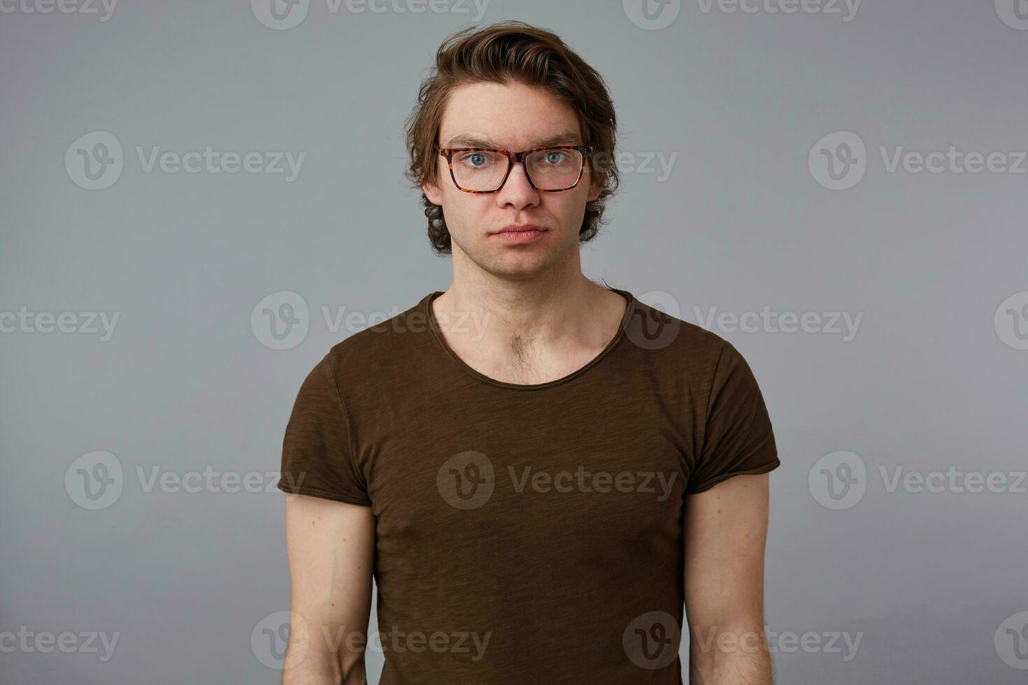 foto do jovem sério cara com óculos desgasta dentro em branco camiseta, carrinhos sobre cinzento fundo e parece às a Câmera sem emoções.