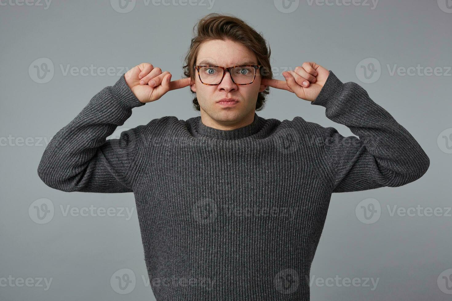 retrato do jovem Bravo homem com óculos desgasta dentro cinzento suéter, carrinhos sobre cinzento fundo, não quer para ouvir, perturbado de ruído e cobre orelhas com dedos. foto