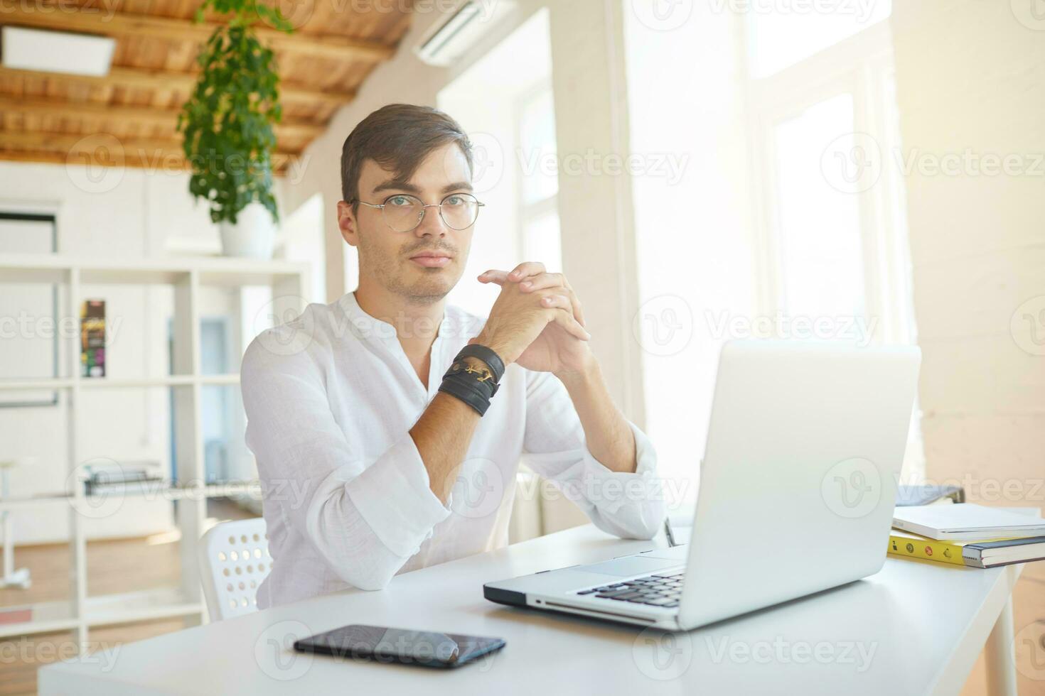 interior tiro do sério concentrado jovem homem de negocios desgasta branco camisa e óculos usando computador portátil e Móvel telefone sentado e pensando às a mesa dentro escritório foto