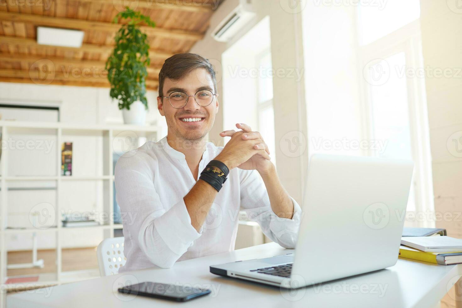 retrato do alegre confiante jovem homem de negocios desgasta branco camisa e óculos usando computador portátil e Smartphone sentado às a mesa dentro escritório foto