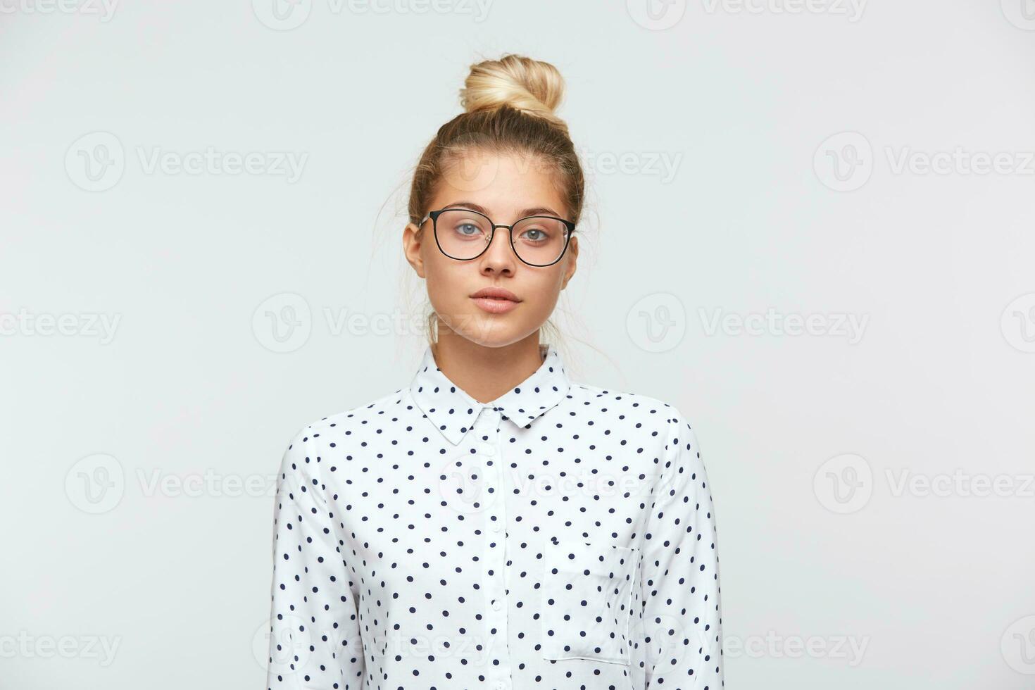 fechar-se do sério lindo jovem mulher com pão desgasta polca ponto camisa e óculos sente infeliz e parece para a Câmera isolado sobre branco fundo foto