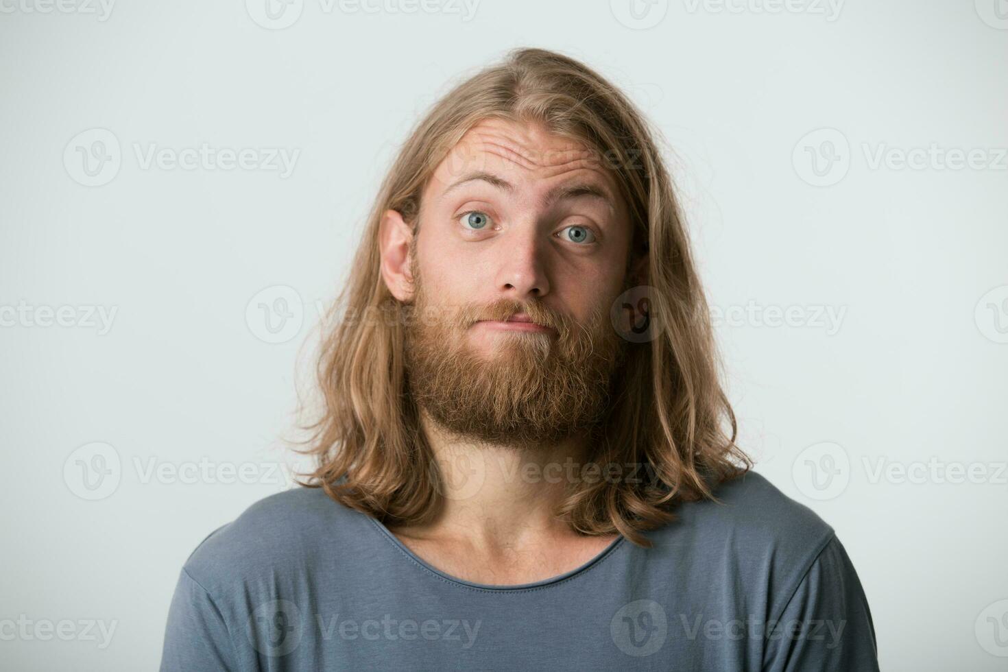retrato do depressivo bonito barbudo jovem homem com Loiras grandes cabelo desgasta cinzento t camisa sente triste e infeliz isolado sobre branco fundo foto