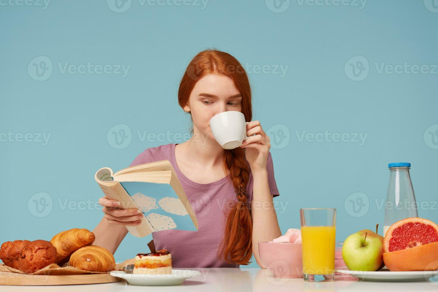cutelo ruivo menina com trançado cabelo, sentado às uma mesa, bebidas a partir de branco copo delicioso chá, tem café da manhã lendo livro. em mesa cozimento produtos e fresco Comida escala azul parede foto