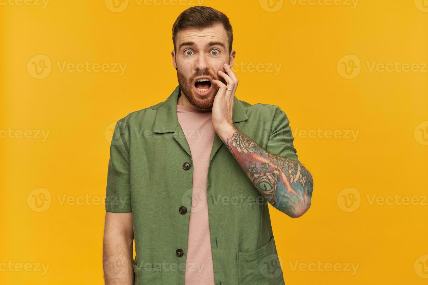 barbudo atordoado cara, descontentamento homem com morena cabelo. vestindo verde curto mangas jaqueta. tem tatuagem. tocante dele face dentro choque. assistindo às a Câmera, isolado sobre amarelo fundo foto