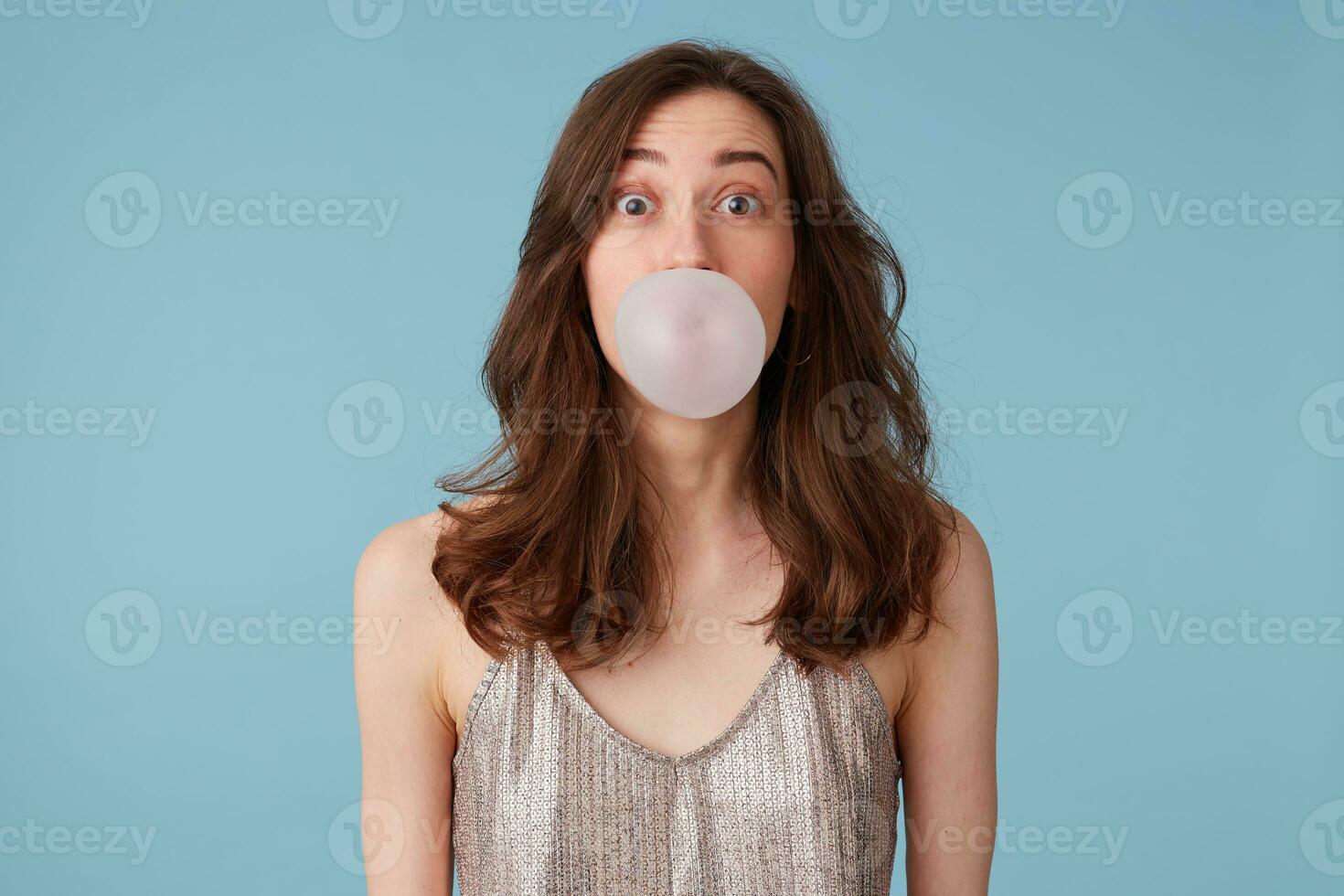 retrato do espantado agradável menina inflar uma bolha, mastigar uma Chiclete, tendo diversão, dentro uma festa vestir, isolado sobre azul fundo foto