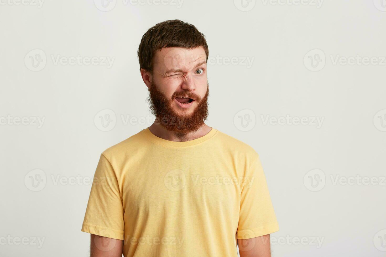 fechar-se do sorridente brincalhão jovem homem hipster com barba desgasta amarelo t camisa sente feliz e piscando isolado sobre branco fundo foto