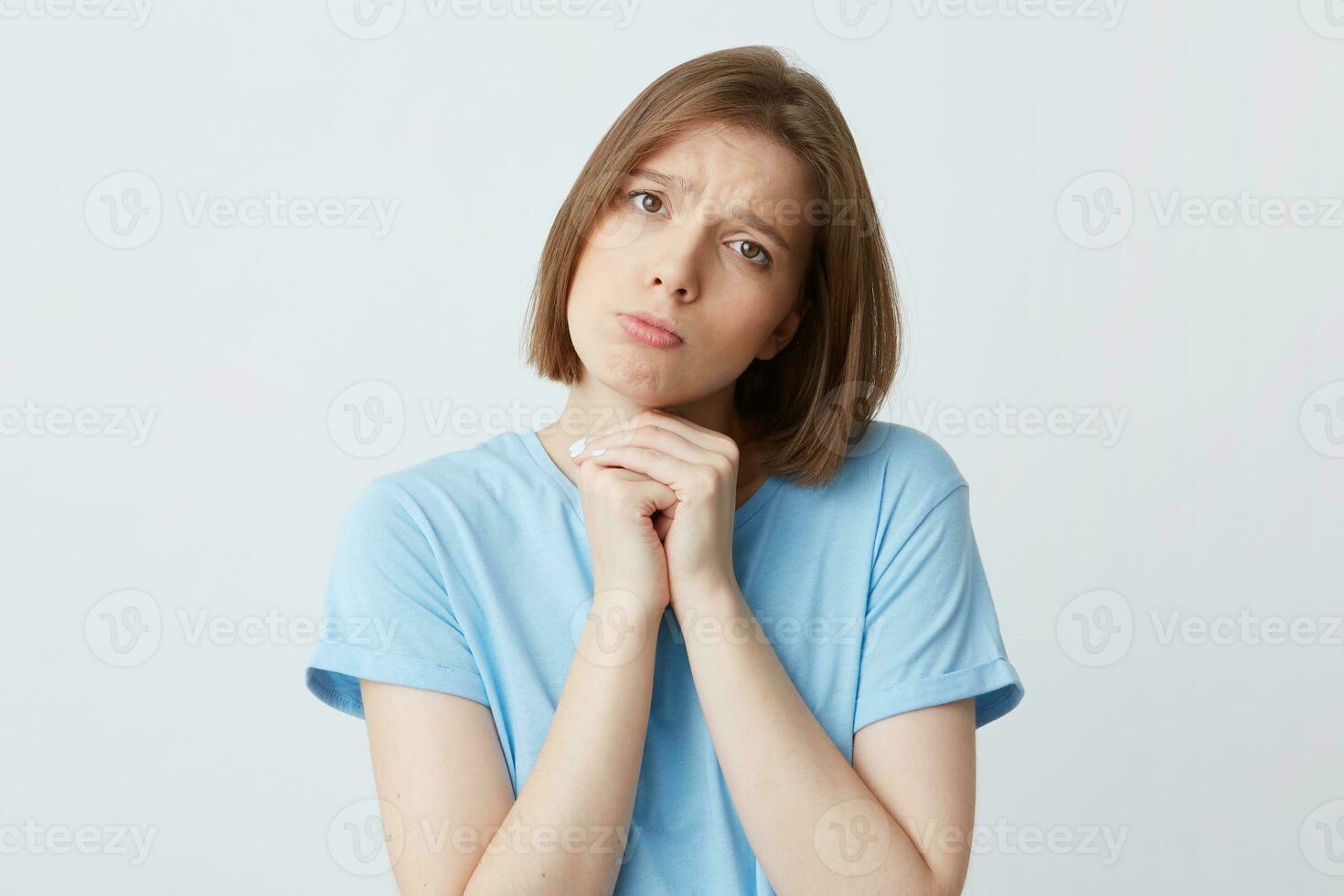 retrato do triste preocupado jovem mulher dentro azul t camisa guardada mãos debaixo dela queixo e sente Desculpe isolado sobre branco fundo parece diretamente dentro Câmera foto
