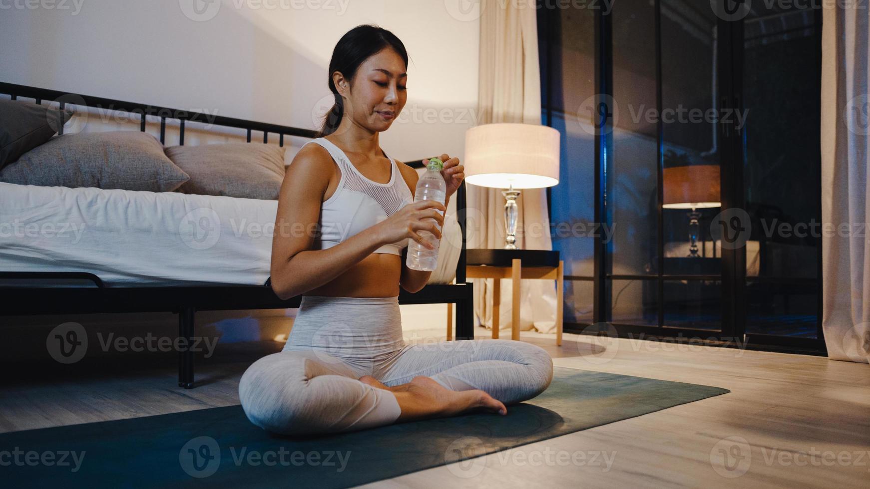 jovem asiática fazendo exercícios de ioga malhando e bebendo água pura na sala de estar em casa à noite. atividade de esporte e recreação, distanciamento social, quarentena para o conceito de prevenção do vírus corona. foto