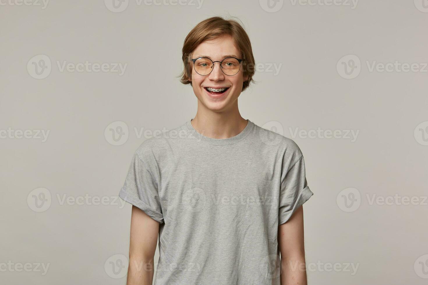 feliz olhando homem, bonito cara com loiro cabelo. vestindo cinzento camiseta, óculos e tem aparelho ortodôntico. pessoas e emoção conceito. assistindo e amplamente sorridente às a Câmera isolado sobre cinzento fundo foto