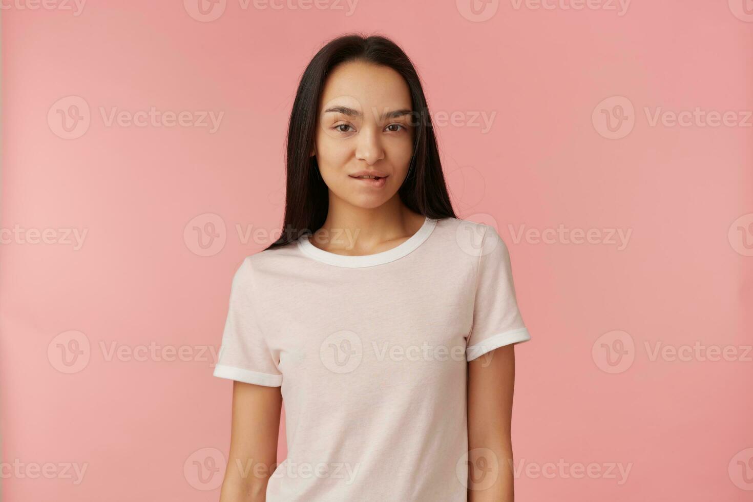 agradável olhando mulher, lindo menina com grandes Preto cabelo. vestindo branco camiseta. pessoas e emoção conceito. mordidas uma lábio e assistindo provocante às a Câmera isolado sobre pastel Rosa fundo foto