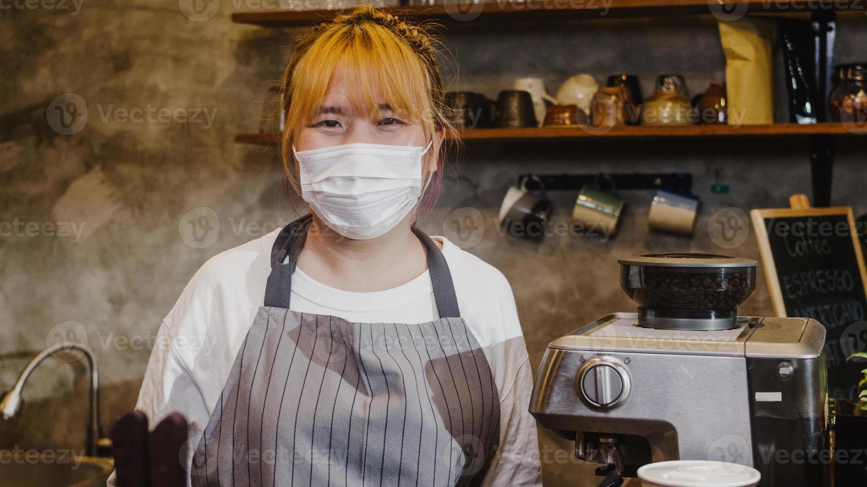 retrato jovem garçonete asiática usar máscara médica sentindo sorriso feliz esperando por clientes após o bloqueio no café urbano. proprietário de uma pequena empresa, comida e bebida, conceito de reabertura de negócios. foto