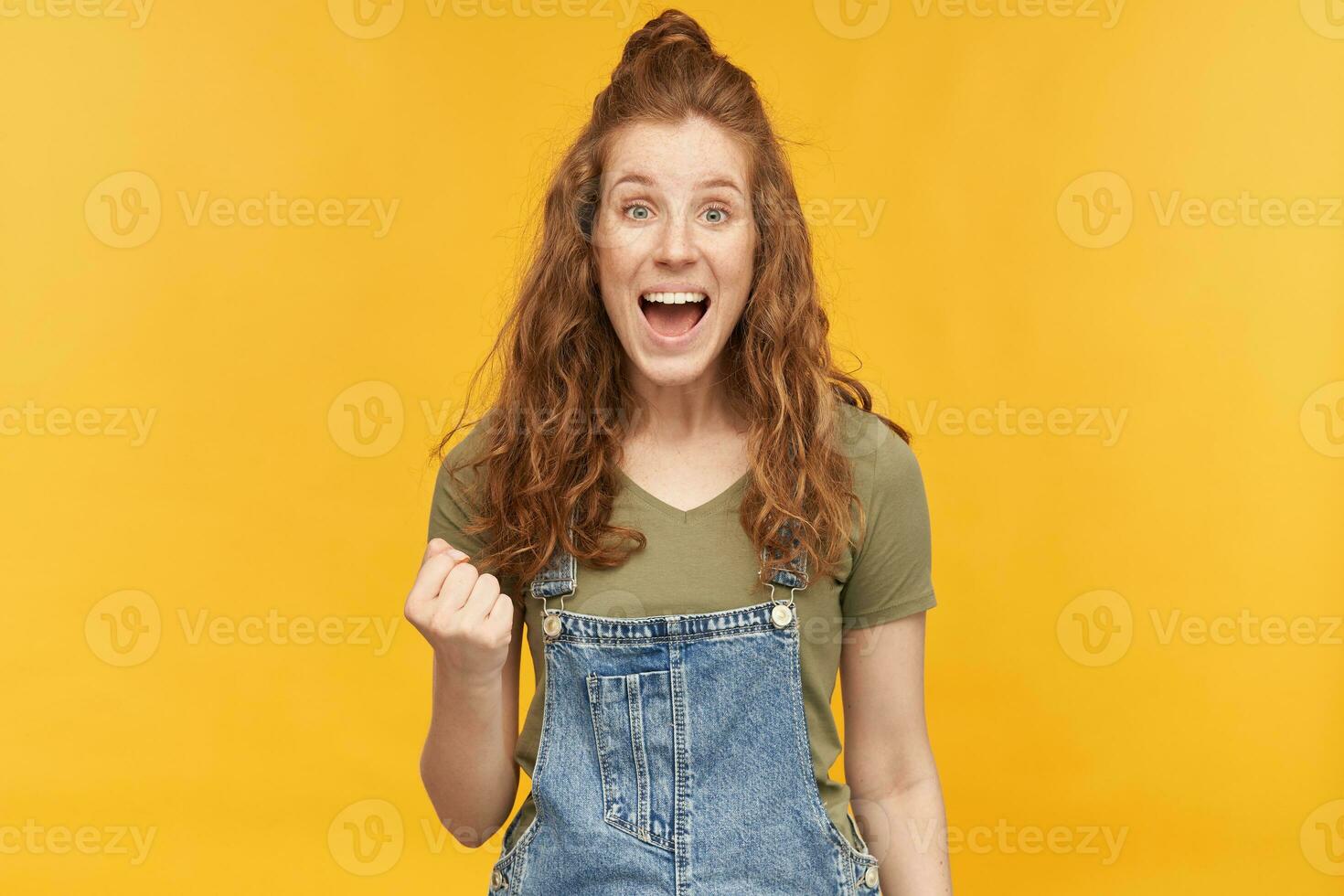 interior tiro do jovem gengibre fêmea desgasta azul jeans macacão gritando, encarando para dentro Câmera e mostra vitória placa com dela mão. isolado sobre amarelo fundo foto