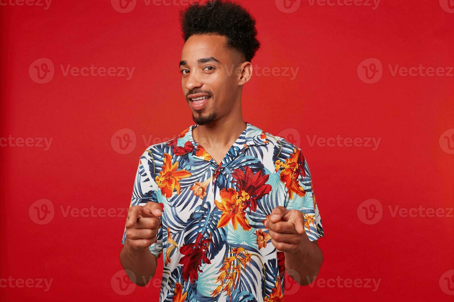 jovem positivo africano americano homem desgasta dentro havaiano camisa, parece às a Câmera e amplamente sorrisos, mostra dedos para a Câmera e diz você legal carrinhos sobre vermelho fundo. foto