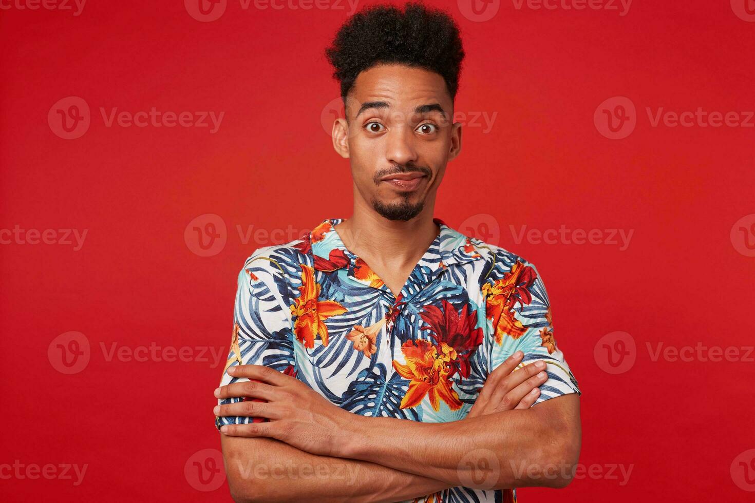 jovem africano americano cara desgasta dentro havaiano camisa, olhando às a Câmera com perguntou-se e mal-entendido, carrinhos sobre vermelho fundo com cruzado braços. foto