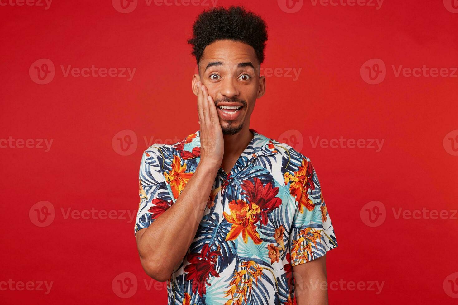 retrato do jovem feliz espantado africano americano cara, desgasta dentro havaiano camisa, parece às a Câmera com surpreso expressão toques bochecha, carrinhos sobre vermelho fundo. foto