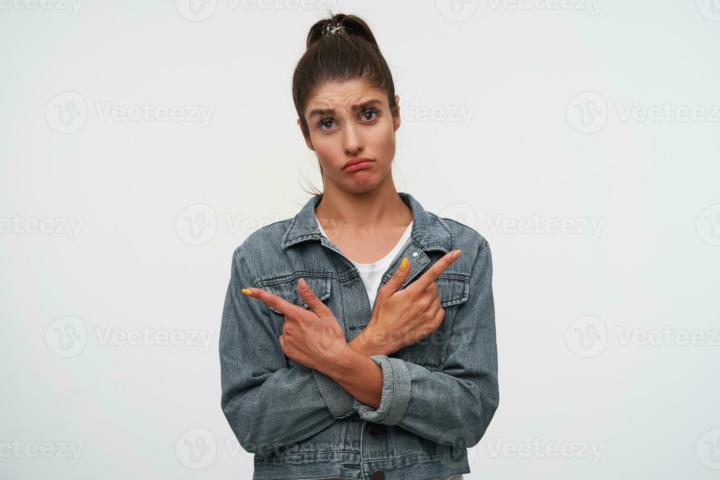 jovem olhar severo morena senhora desgasta dentro branco camiseta e jeans jaquetas, parece às a Câmera com infeliz expressão, pontos com dedos dentro diferente instruções, carrinhos sobre branco fundo. foto