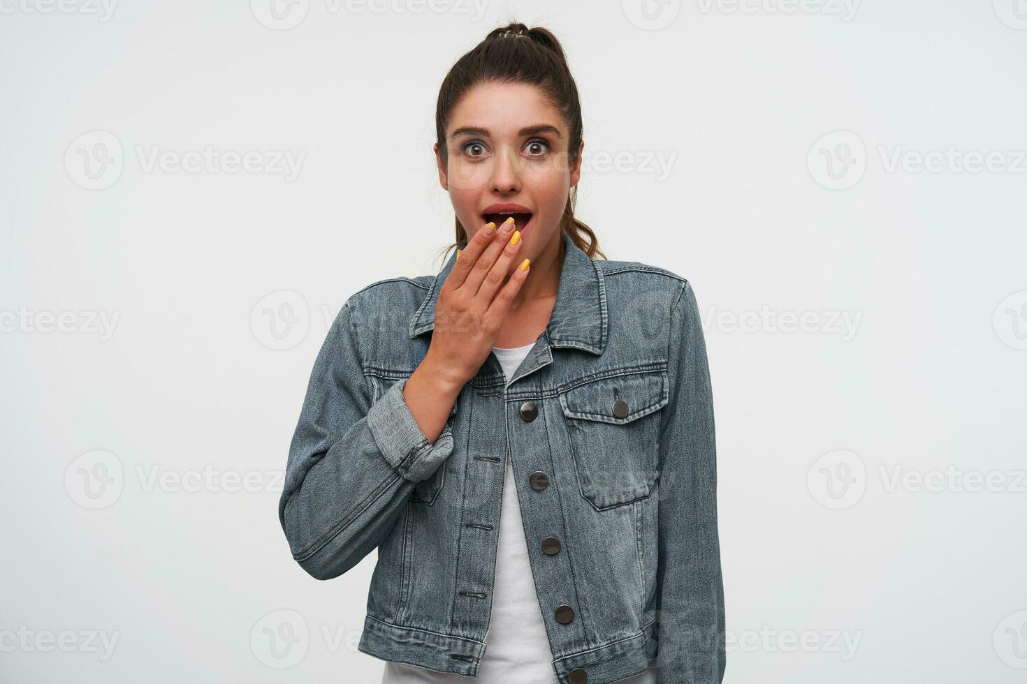 jovem feliz espantado morena senhora dentro branco camiseta e jeans jaquetas, parece às a Câmera com Largo aberto boca e olhos dentro surpreso expressão, coberto boca com Palma, carrinhos sobre branco fundo. foto