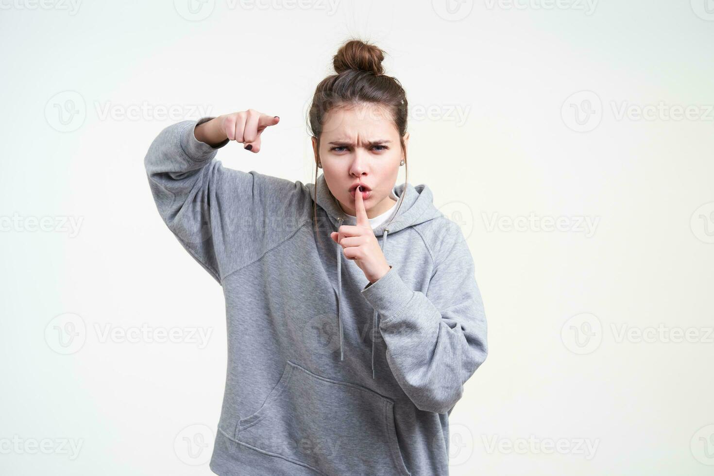 sombrio jovem Castanho cabelos mulher com natural Maquiagem guardando indicador em dela boca enquanto mostrando severamente às Câmera, em pé contra branco fundo foto