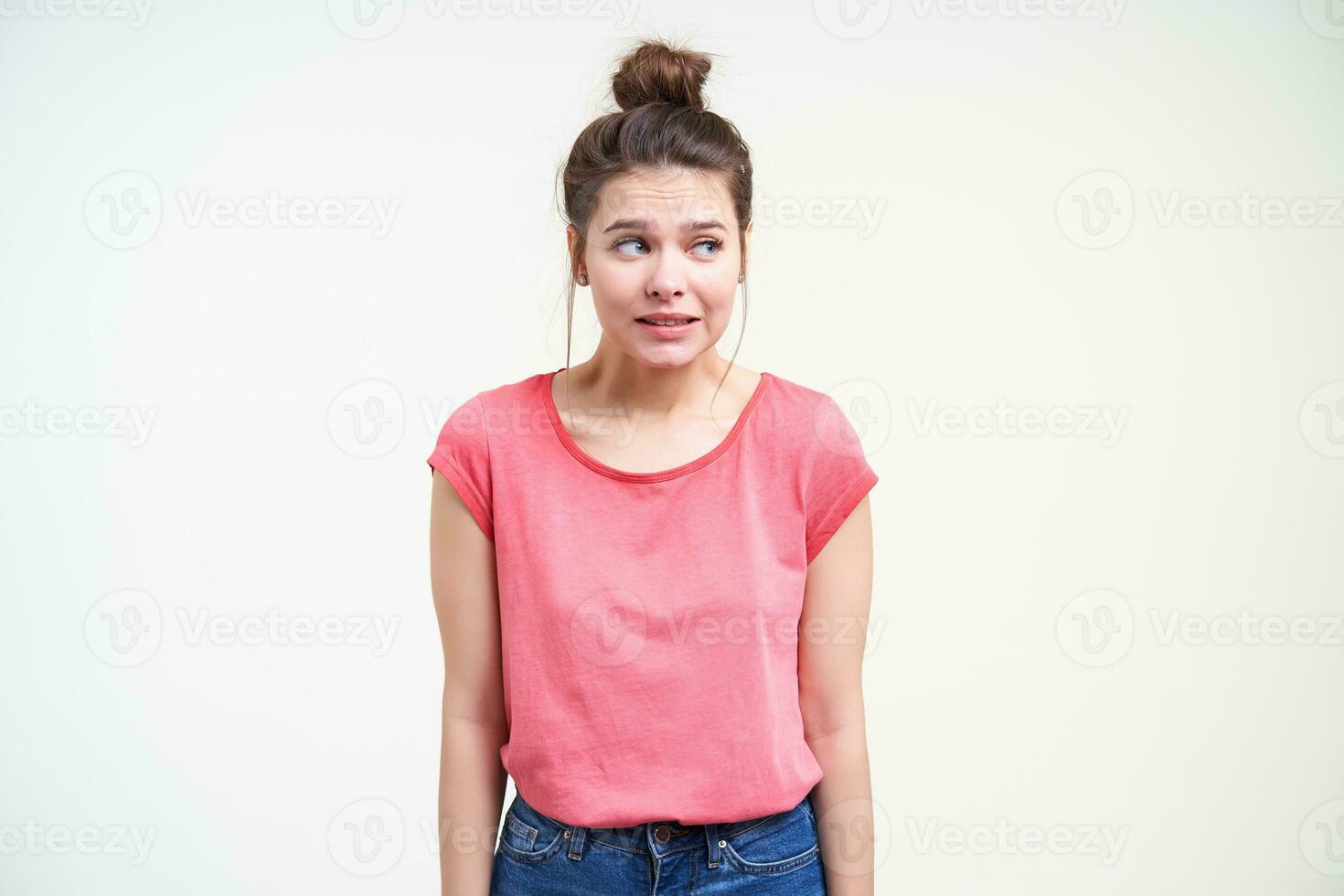 estúdio foto do jovem intrigado Castanho cabelos fêmea olhando confusamente a parte, de lado e guardando mãos baixa enquanto em pé sobre branco fundo dentro Rosa camiseta