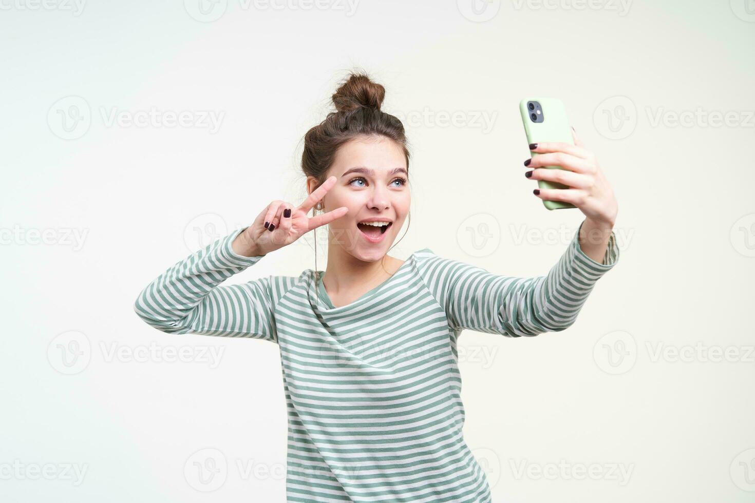 alegre jovem de olhos azuis adorável morena fêmea com pão Penteado mostrando Paz placa com elevado mão enquanto levando selfie com dela Smartphone, isolado sobre branco fundo foto