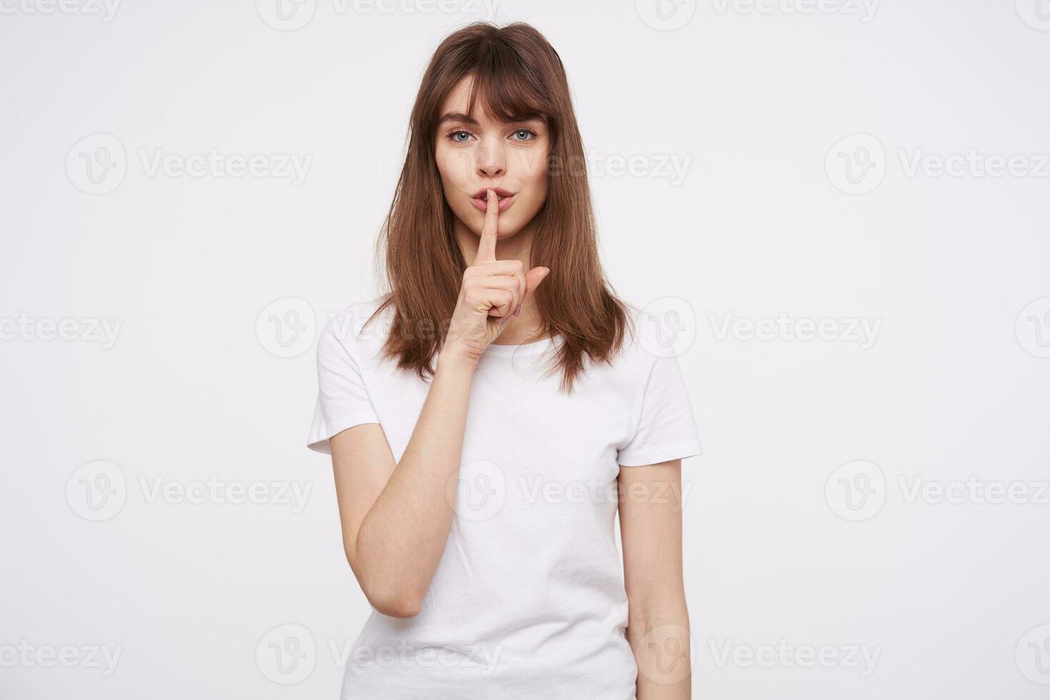 retrato do lindo jovem calma Castanho cabelos mulher com natural Maquiagem segurando indicador em dela lábios enquanto em pé sobre branco plano de fundo, Perguntando para manter segredo foto