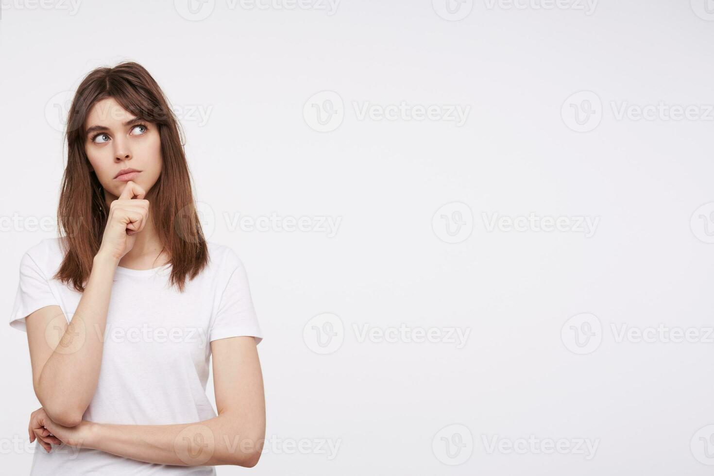 pensativo jovem bonita morena mulher segurando dela queixo com elevado mão enquanto olhando pensativamente para cima, guardando dela lábios guardada enquanto em pé sobre branco fundo foto