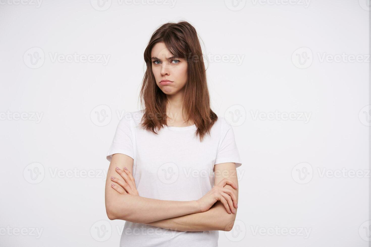 forte jovem de olhos azuis morena mulher com natural Maquiagem franzindo a testa dela sobrancelhas enquanto olhando a sério às Câmera, guardando mãos guardada enquanto posando sobre branco fundo foto