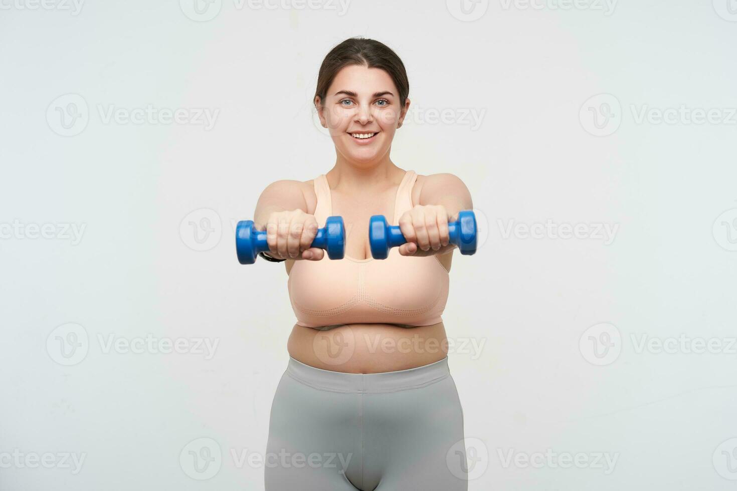 estúdio tiro do jovem satisfeito de grandes dimensões fêmea fazer estático exercícios com peso e sorridente de bom grado às Câmera, trabalhando Fora dela corpo enquanto posando sobre branco fundo foto