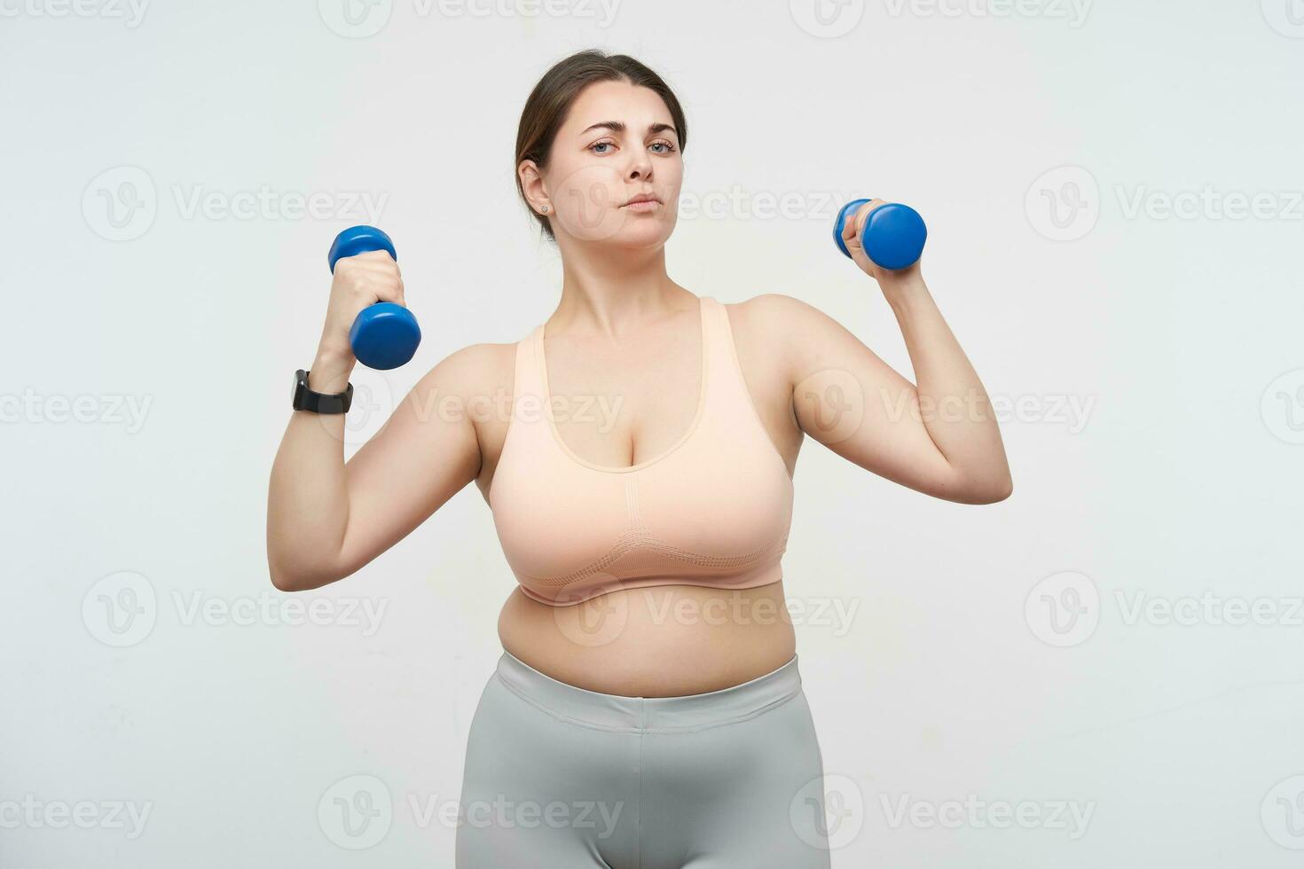 interior foto do jovem morena gordo senhora com casual Penteado olhando a sério às Câmera enquanto trabalhando Fora com halteres, isolado sobre branco fundo