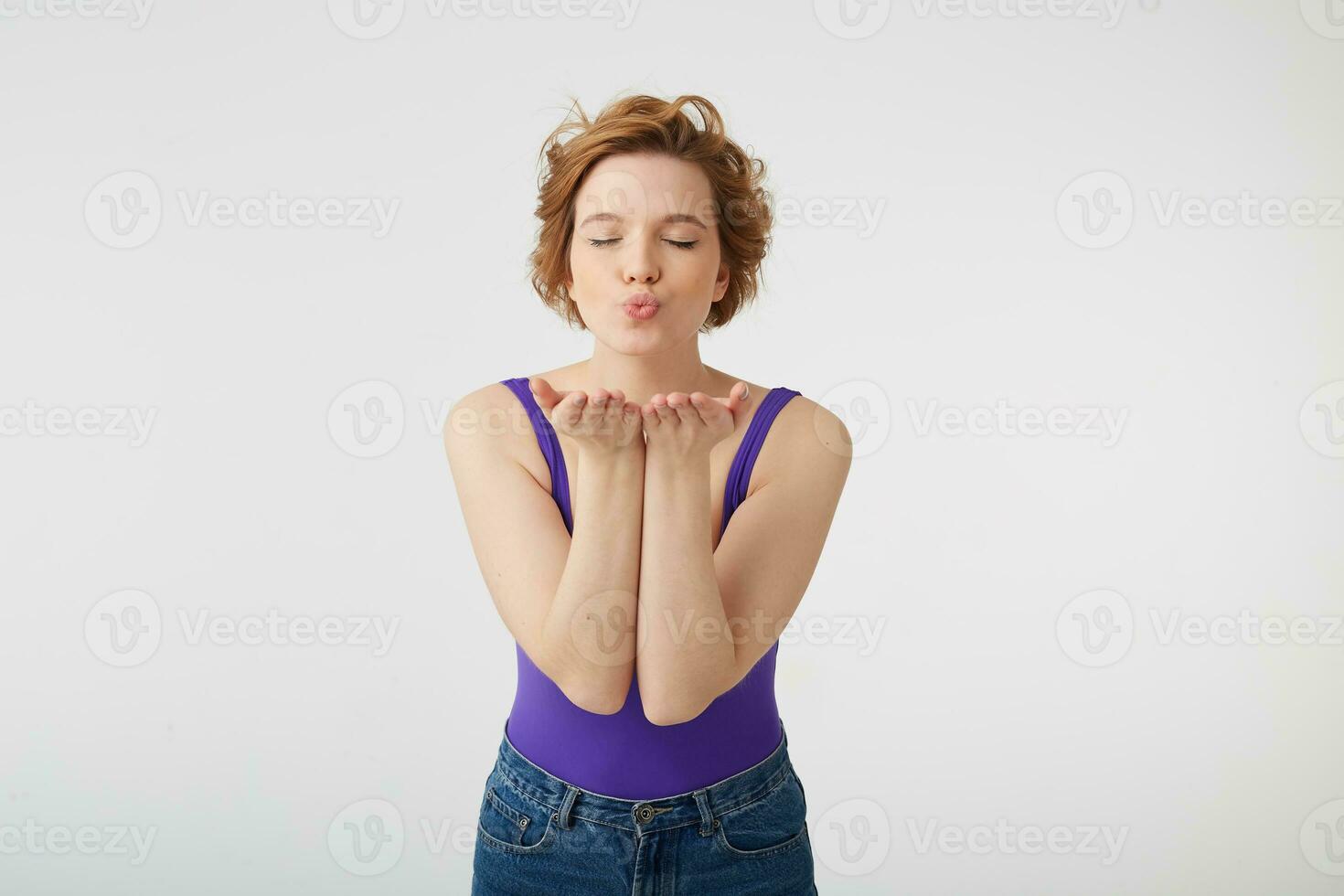 retrato do jovem agradável cabelo curto menina desgasta dentro roxa camisa, mostra ar beijo, sobressai Palmeiras às Câmera, expressa amor para alguém em distância, carrinhos com fechadas olhos sobre branco fundo. foto