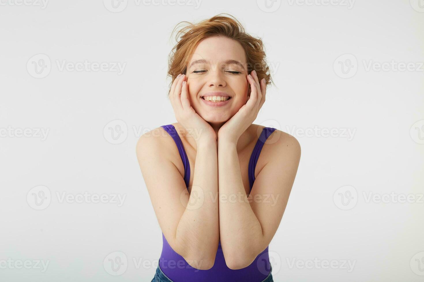 fechar acima do jovem agradável cabelo curto senhora desgasta dentro roxa camisa e jeans, fecha olhos e sonhos sobre Novo vestir, toques dele bochechas e sobre branco fundo. foto