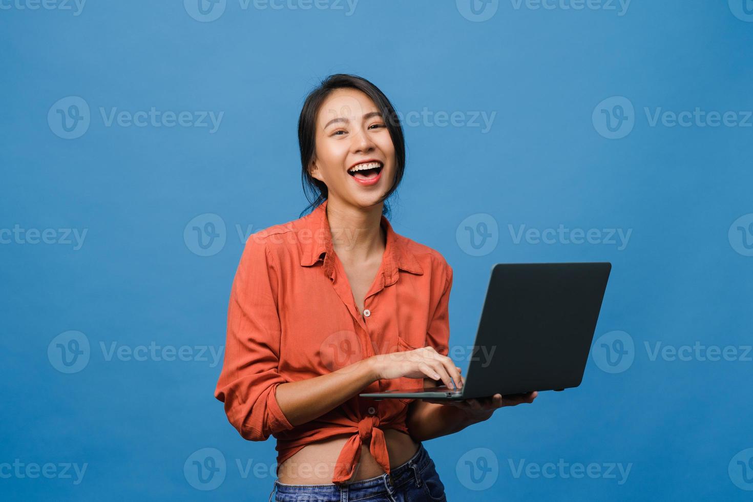 surpreendeu a jovem asiática usando laptop com expressão positiva, sorriso largo, vestido com roupas casuais e olhando para a câmera sobre fundo azul. feliz adorável feliz mulher alegra sucesso. foto